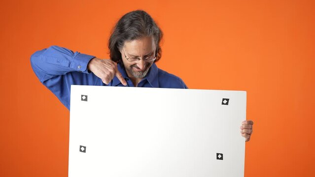 Happy smiling mature man in blue shirt isolated on orange background holding white blank copy board sign with tacking points and place for text.