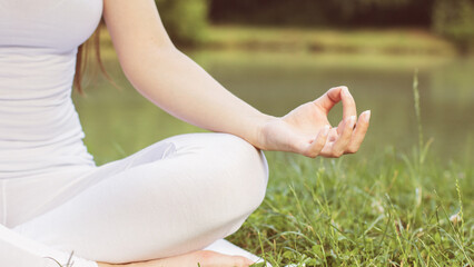 Closeup view of female hand during meditation at lake on summer. Girl wearing white activewear while practicing yoga. Balance and harmony. Healthy lifestyle concept. Nature landscape in background.