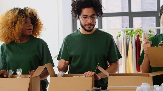 charity, donation and volunteering concept - international group of happy smiling volunteers packing food in boxes according to list on clipboard at distribution or refugee assistance center