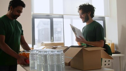 charity, donation and volunteering concept - male volunteers with clipboard packing food and drinks in boxes over international group of people at distribution or refugee assistance center