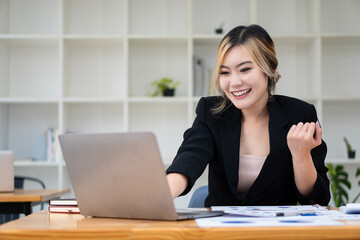 Beautiful Asian businesswoman analyzing chart using calculator, laptop at the office with papers and holding pen, sitting on the chair at modern home studio. Mobile worker contacting customers