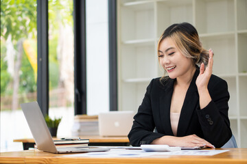 Beautiful Asian businesswoman analyzing chart using calculator, laptop at the office with papers and holding pen, sitting on the chair at modern home studio. Mobile worker contacting customers