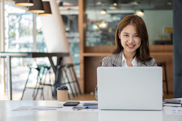 Beautiful Asian businesswoman analyzing chart using calculator, laptop at the office with papers and holding pen, sitting on the chair at modern home studio. Mobile worker contacting customers