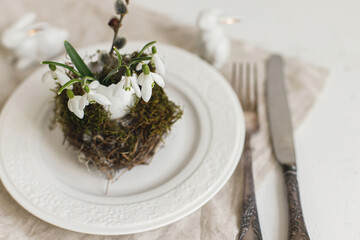 Stylish Easter table setting. Natural egg with snowdrops in nest on modern plate, napkin, cutlery, bunny on white wood. Modern Easter table rustic decoration