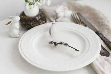 Stylish Easter table setting. Pussy willow branch, feather on modern plate, napkin, cutlery, bunny and egg with snowdrops on white wooden table. Modern Easter table decoration