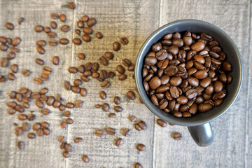 on a wooden table with scattered coffee beans is a dark mug filled with roasted whole coffee beans. top view