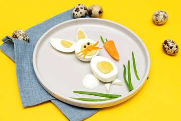 Easter funny creative healthy breakfast lunch food idea for kids, children.Bunny, rabbit made from boiled chicken eggs,peeled carrots, greens on plate yellow table background.Top view Flat lay