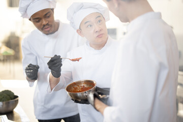 Three chef cooks with different ethnicities tasting sauce with a spoon while cooking in the kitchen. Teamwork and cooking delicious food at restaurant