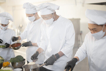 Multiracial team of chef cooks in white uniform cooking together in the kitchen. Asian, latin and european guys cooking together. Cooks wearing face masks and protective gloves 