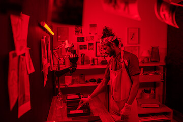 Creative young Indian man wearing apron and gloves standing in darkroom at home developing photo prints