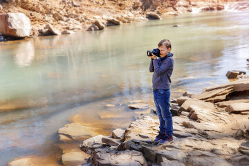 Photographer boy near a mountain river standing on the rock. Kid enjoying the moment of the beautiful blue water in sunny day. Boy is holding photo camera and taking photo of the view.