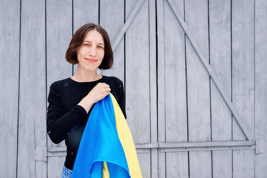 Young Girl Holding A Blue And Yellow State Flag Of Ukraine In Her Right Hand Over A Heart On A Background Of Gray Wooden Fence. A Woman In A Black Jacket And Jeans Stands On The Street Under The Snow