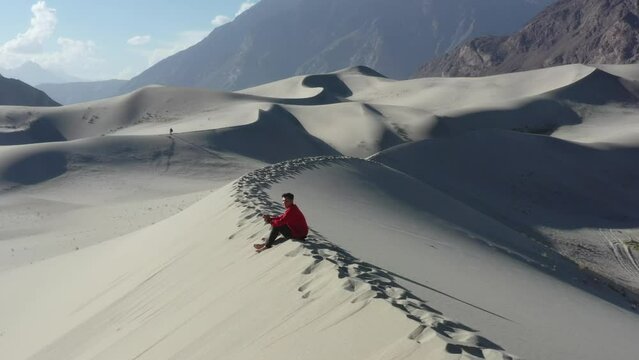 young male piloting a drone in a red jacket sitting on top of sand dunes in the cold desert of skardu pakistan during a sunny day with large mountains in the distance