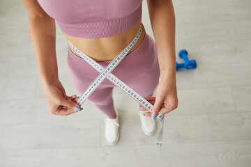 Slim young girl holding tape measure on her thin waist. Unrecognizable sporty woman showing her weightloss achievement after home workouts. Cropped close up, closeup. Beauty and weight loss concept - obrazy, fototapety, plakaty