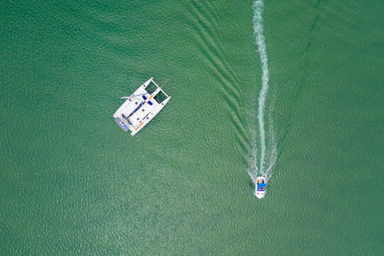 Looking down on a single catamaran and a small boat speeding past leaving a wake in the calm water