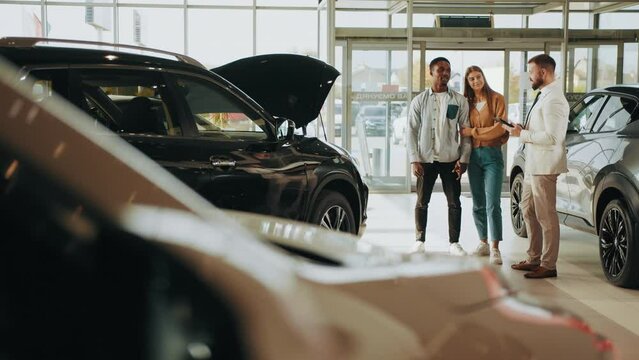 Competent car dealer using digital tablet for work with clients at showroom. Multiracial couple talking with manager while buying car at dealership center. Translation: Thank you.