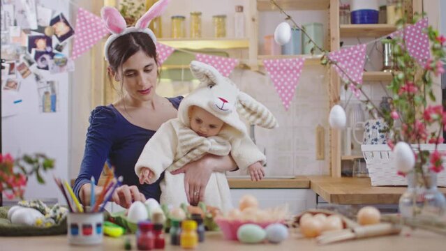 Mother with baby decorating easter eggs. Baby at rabbit costume and mother with bunny ears at the kitchen celebrating Easter.