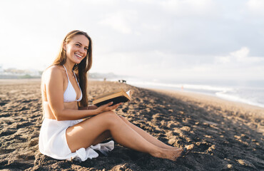 Fototapeta na wymiar Portrait of funny Caucaisan woman with literature book in hands laughing at sandy beach near Caribbean sea smiling at camera during leisure, joyful female reader with best seller enjoying weekend