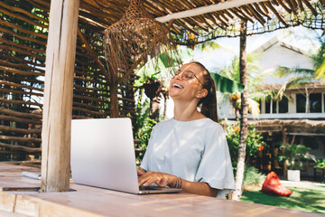 Joyful graphic designer in eyeglasses using netbook computer during remote working outdoors, cheerful female copywriter with laptop laughing and rejoicing - digital nomad and freelance lifestyle