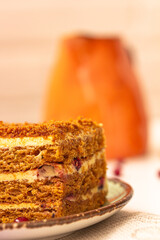 Homemade layered honey cake with cream and berries on the plate, a bright orange creamer on beige ...