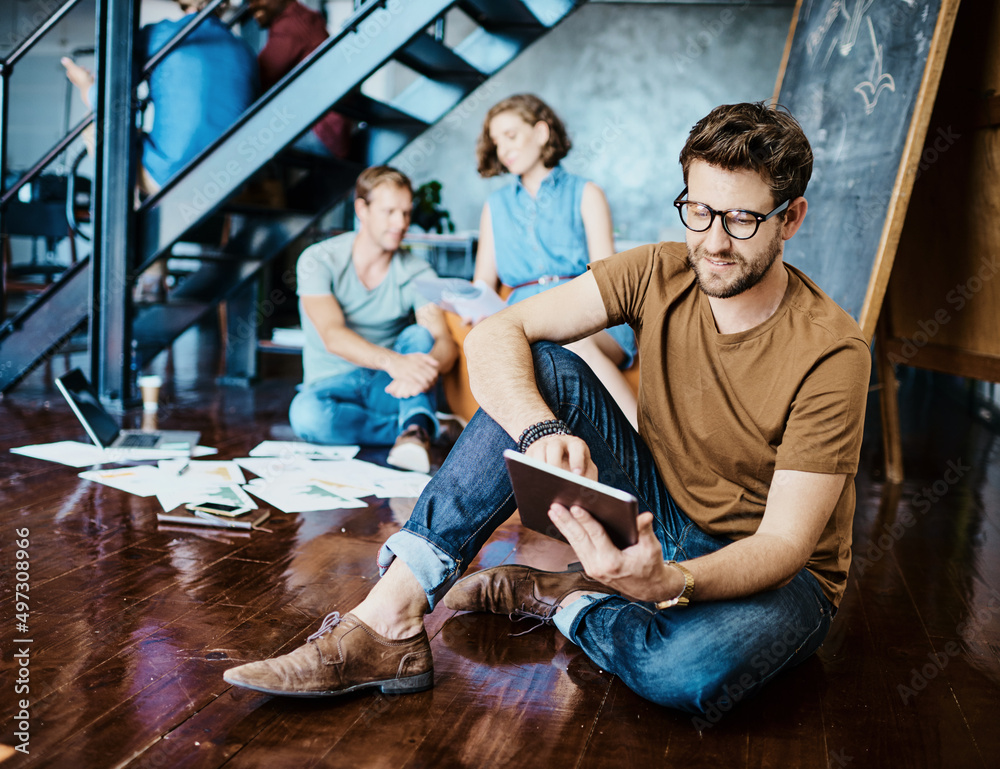 Poster Using smart tools to gain a competitive edge. Shot of a young designer working on a digital tablet with his colleagues in the background.