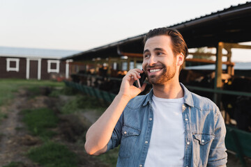 happy farmer looking away while talking on smartphone near blurred cowshed.