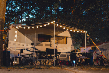 Campervans are parked in forest. Camping equipment. Lights, table and chairs. Night time.