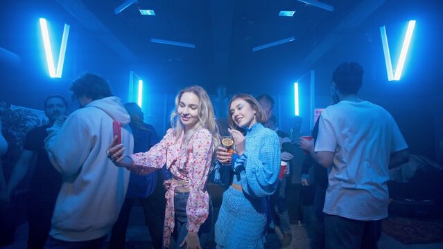 Two girls with a cocktail in their hands are dancing, a crowd of people is dancing around the girls on the dance floor of a nightclub under the light of colored spotlights. Girls take selfies on the