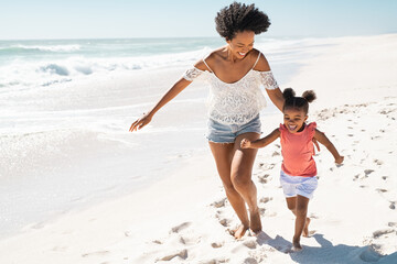 Playful cute little daughter running on beach with young black mother