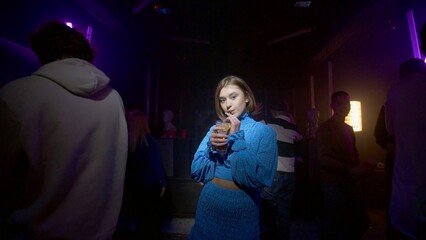 A cute girl with a cocktail in her hands is dancing and looking at the camera, around the girl a crowd of people is dancing on the dance floor of a nightclub under the light of colored spotlights