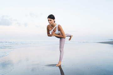 Concentrated and flexible female trainer in sportive tracksuit stretching body muscles and doing warm up exercises at coastline sand, healthy Caucasian woman 20s keeping vitality and wellness