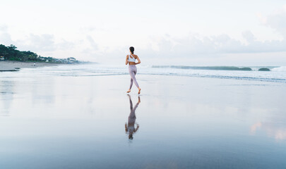 Back view of woman in stylish tracksuit running at seashore for keeping perfect slim figure, barefoot energetic female in sportive clothes have cardio training while jogging at coastline beach