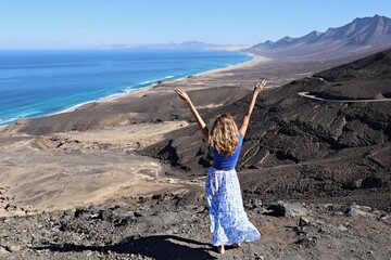 Radosna turystka z rękami do góry stojąca tyłem i podziwiająca piękny widok na plaże Cofete na Fuerteventura - obrazy, fototapety, plakaty