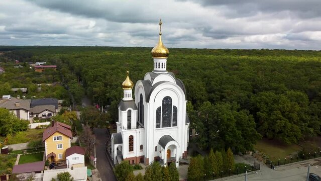 White Cathedral with golden dome aerial fly clockwise around view with dramatic clouds. Kharkiv city Pavlovo Pole district, view on Nauky ave, christian church and green vivid forest