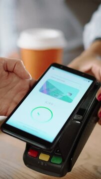 Vertical Footage of a Close Up of a Hand Holding a Bank Terminal Used for NFC Payment Technology on Smartphone. Customer Uses Mobile to Pay for Take Away Latte to Barista in a Coffee Shop.