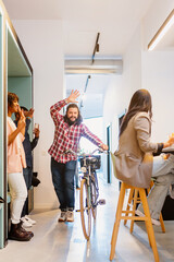 Caucasian middle-aged man entering an office with his bicycle in hand and greeting his co-workers. Multiracial group of workers in an office.
