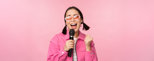 Happy beautiful asian girl singing with mic, using microphone, enjoying karaoke, posing against...