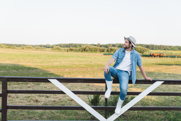 full length view of farmer in denim clothes and brim hat sitting on wooden fence on farmland.