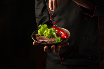 A professional chef sprinkles salt on a sliced beef steak and vegetables in a plate. The concept of serving dishes to order