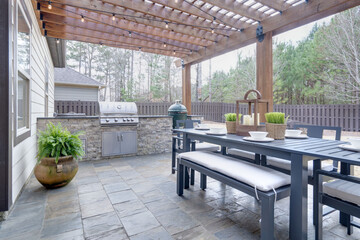 Pretty summer outdoor kitchen with table set and grilling station underneath wooden arbor on stone patio. - obrazy, fototapety, plakaty