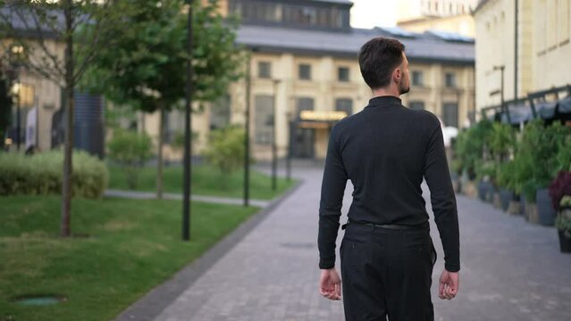 Back view young confident man strolling on city street in the morning. Live camera follows Caucasian successful businessman walking in slow motion looking around. Style and confidence concept