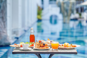Breakfast at European style hotel near swimming pool. Table with delicious food by the beautiful blue pool travel in resort on sunny summer day. Rest and vacation in modern luxury hotel
