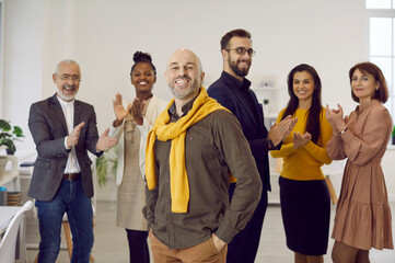 Smiling business team leader. Happy confident successful handsome bald man in casual wear standing hands in pockets, with team of people applauding in background. Recognition at work, success concept