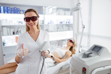 Joyful female beautician with laser device giving thumbs up