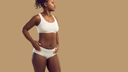 Gorgeous plus size female model posing in underwear. Confident young African American woman wearing white bralette and underpants standing with hands on hips on solid beige color text space background