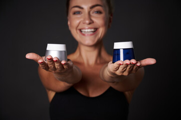 Focus on anti wrinkles moisturizer jars in the hands of a blurred smiling middle aged blonde woman, isolated over black background with copy ad space