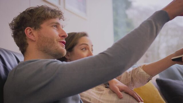 Loving Pregnant Couple Sitting On Sofa At Home Watching TV With Man Lovingly Touching Woman's Bump  - Shot In Slow Motion