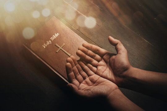A bible on a wooden table illuminated from above. and praying hands concept of faith, hope, trust in God