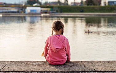 View from behind of sad abandoned little girl with braided hair sitting alone looking at lake...