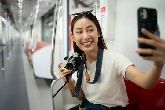 Selective focus of a happy Asian female traveler sitting in a sky train using a mobile taking selfie while the other hand holding a film camera with a blurred train aisle without people in background.